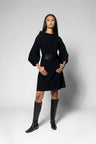 A woman modeling a black long-sleeve mini dress with a wide black belt, paired with black knee-high boots, against a light gray studio background.