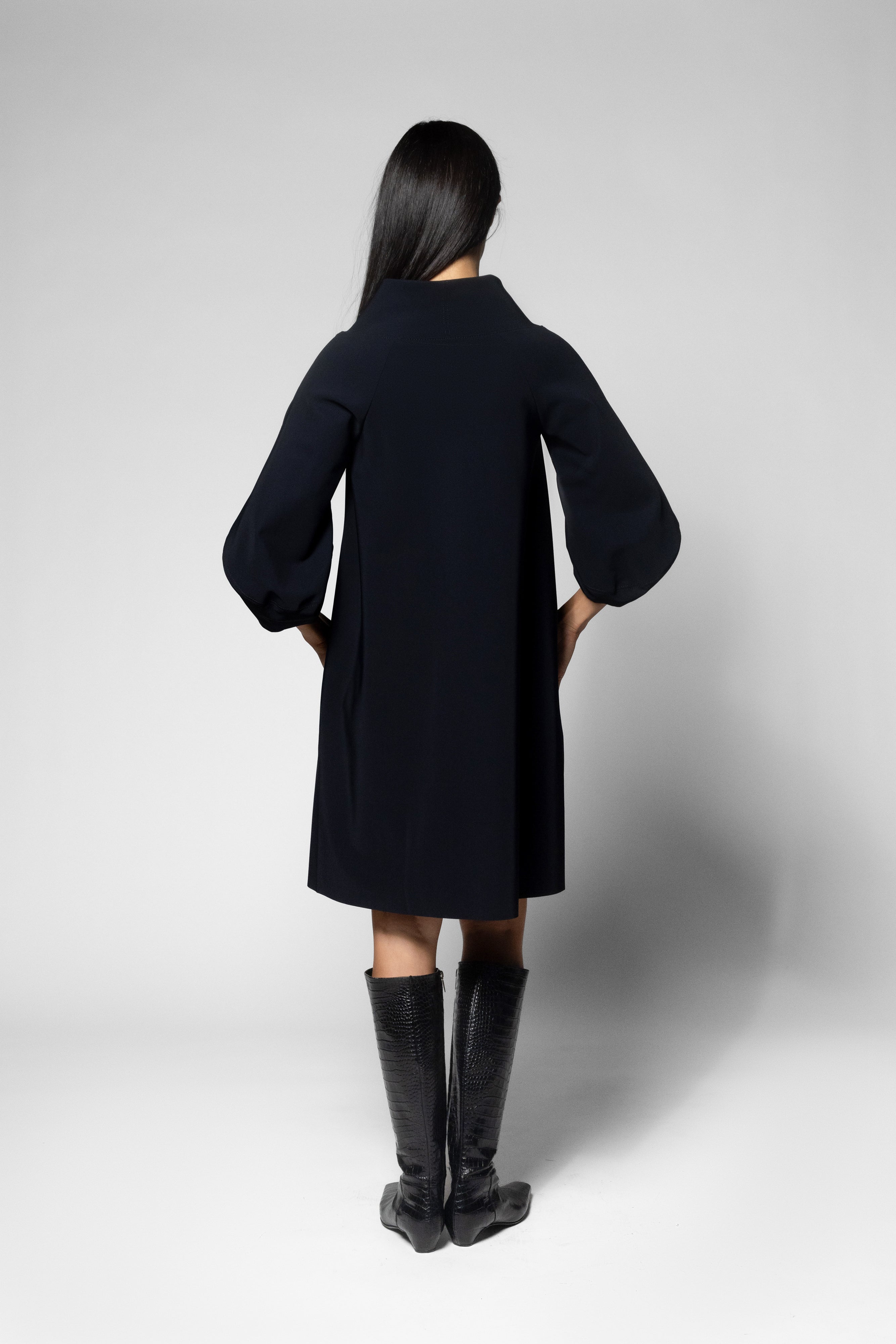 A model viewed from the back wearing a black long-sleeve coat with balloon sleeves and black knee-high boots, set against a plain light gray studio background.
