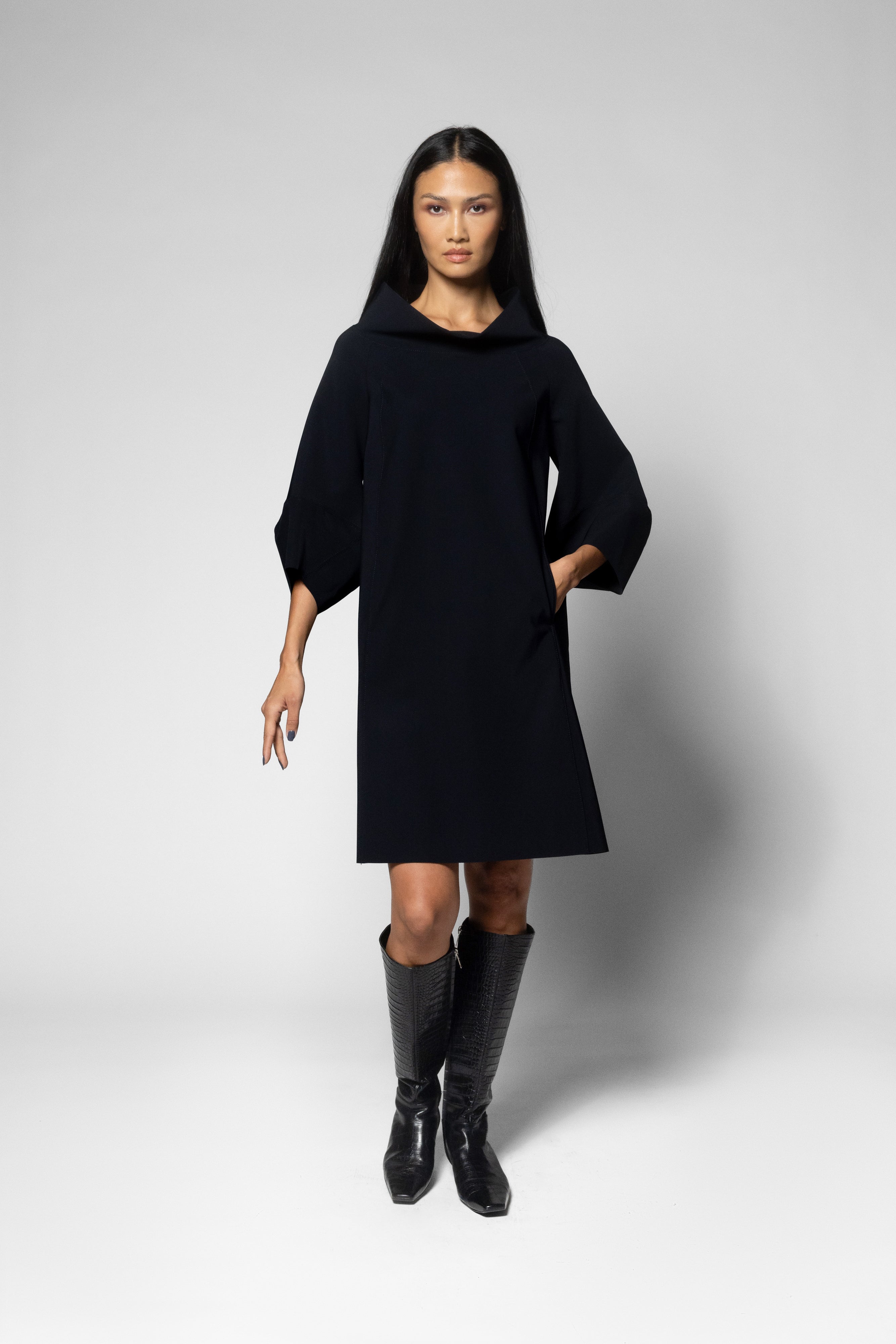 A model wearing a black dress with wide three-quarter sleeves and black knee-high boots, standing against a light gray studio background.