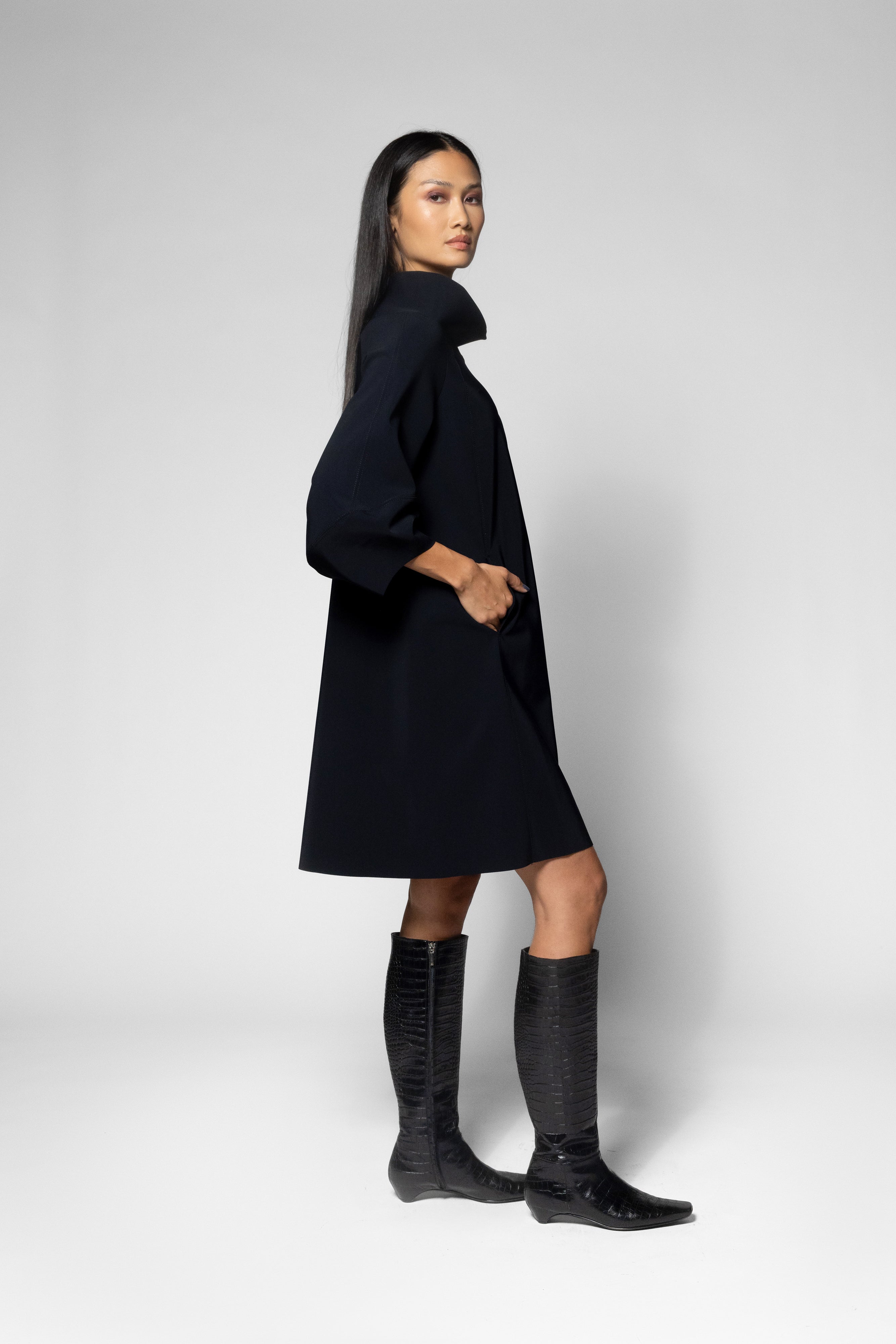 A model wearing a black mid-length dress with a high collar and three-quarter sleeves, paired with black knee-high boots, standing against a light gray studio background.