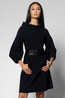 A woman modeling a black dress with a wide collar, balloon sleeves, and a black belt at the waist, photographed against a light gray studio background.
