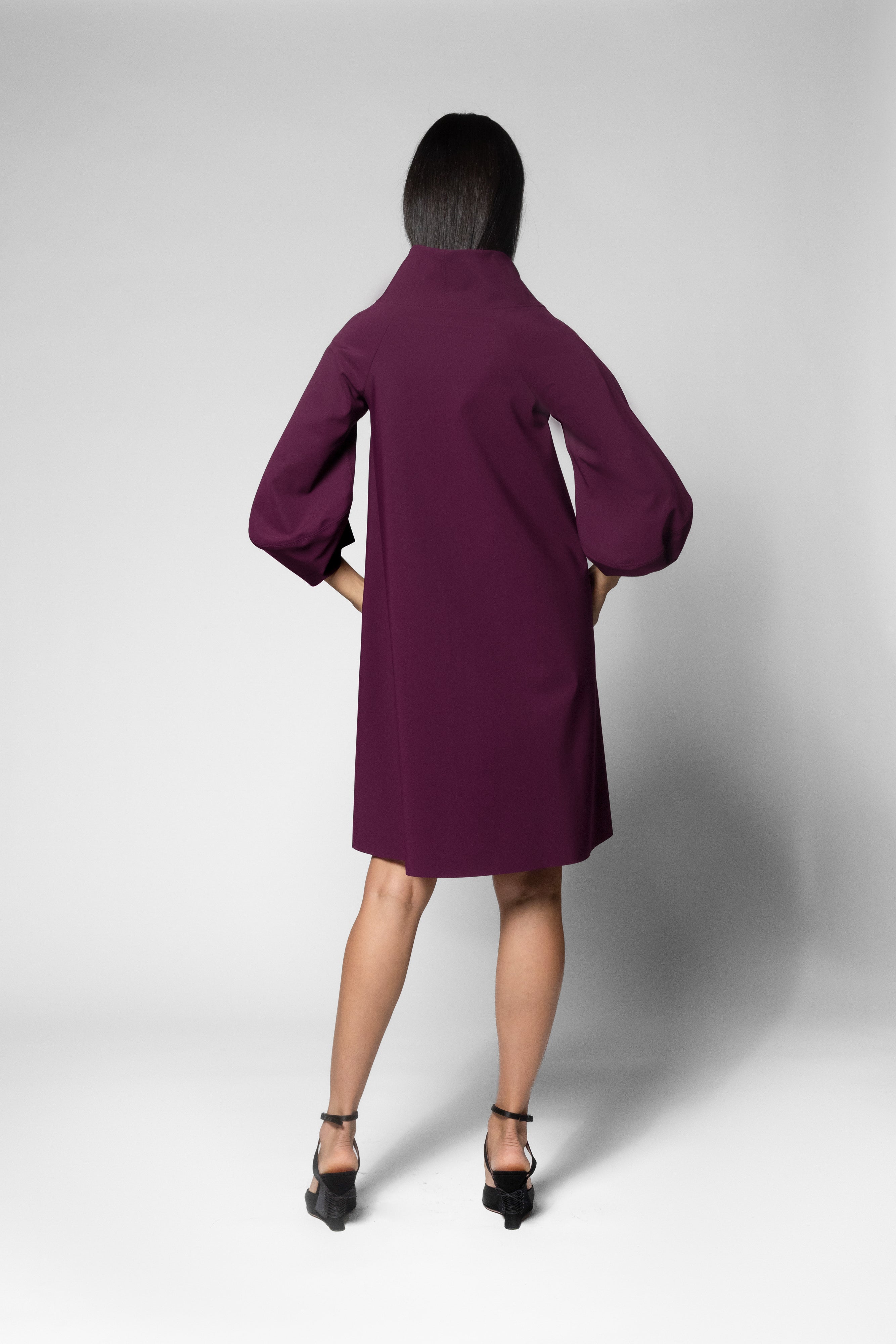 A model viewed from the back wearing a deep purple mid-length dress with voluminous three-quarter sleeves, paired with black ankle-strap heels, against a light gray studio background.