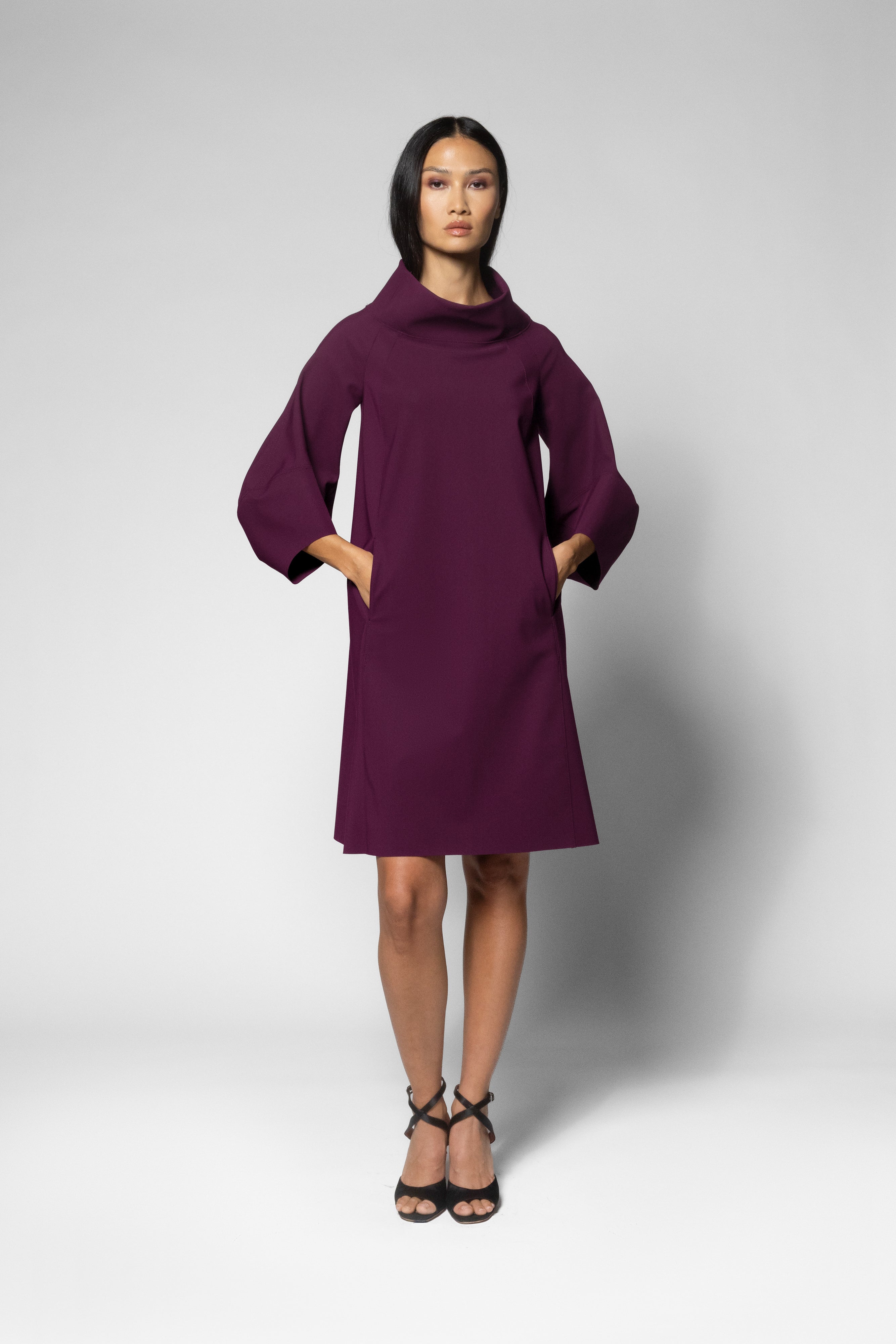 A model wearing a dark purple cowl-neck dress with wide three-quarter sleeves and black strappy high-heeled sandals, standing against a light gray studio background.
