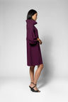A woman modeling a long-sleeved, high-neck, knee-length dress in deep plum purple, shown from a side profile against a light gray studio background.