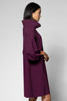 A woman shown in side profile wearing a long, loose-fitting high-neck dress in a deep plum purple, photographed against a plain light gray studio background.