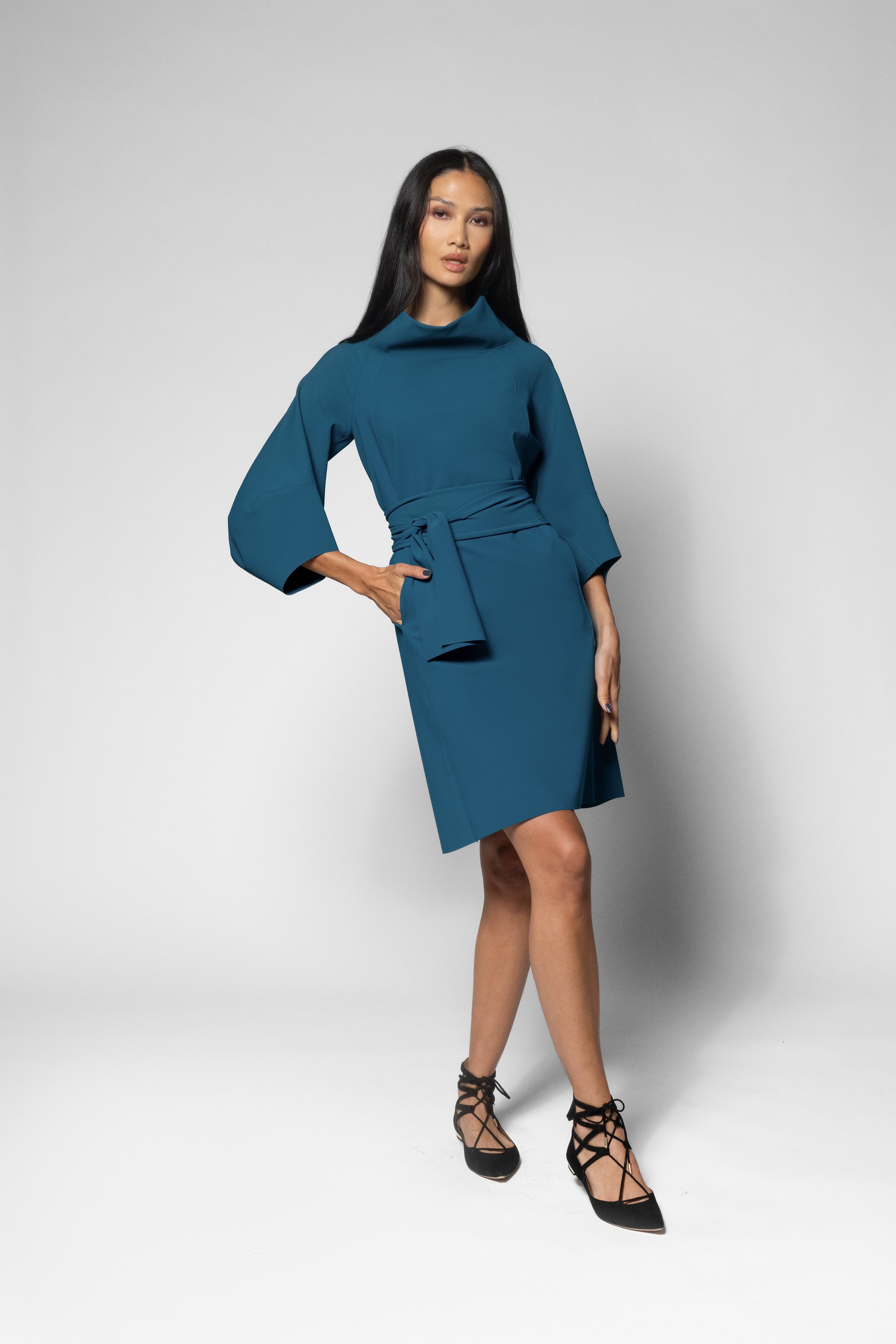 A woman modeling a teal blue wrap-style dress with wide three-quarter sleeves and a matching belt, paired with black lace-up heels, against a light gray studio background.