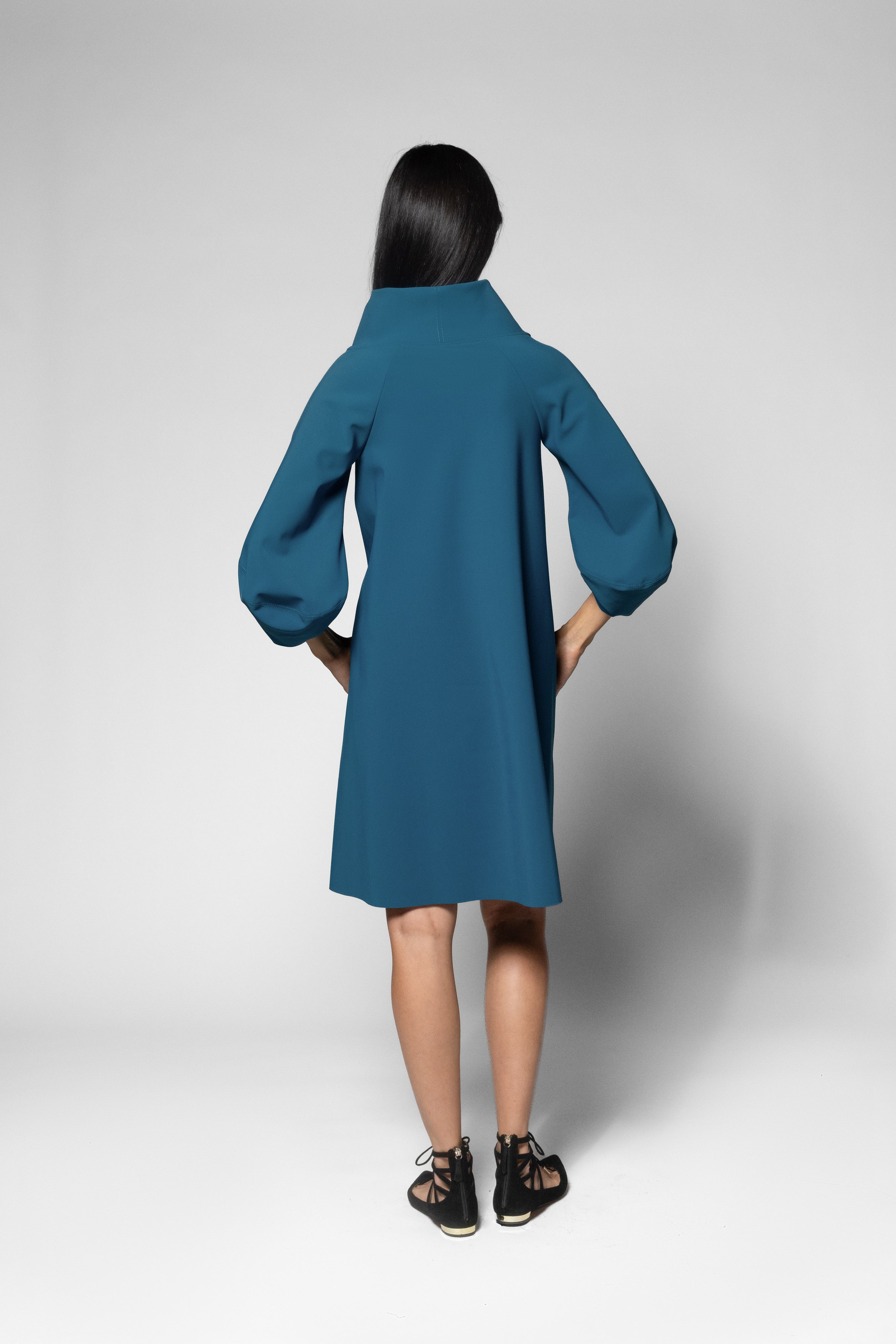 A model viewed from the back wearing a teal blue dress with balloon sleeves and black sandals, standing against a light gray studio background.