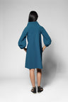 A model viewed from the back wearing a teal blue dress with balloon sleeves and black sandals, standing against a light gray studio background.