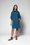 A woman modeling a teal blue cowl-neck shift dress with elbow-length sleeves and side pockets, paired with black lace-up flat sandals, against a light gray studio background.