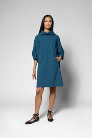 A woman modeling a teal blue cowl-neck shift dress with elbow-length sleeves and side pockets, paired with black lace-up flat sandals, against a light gray studio background.