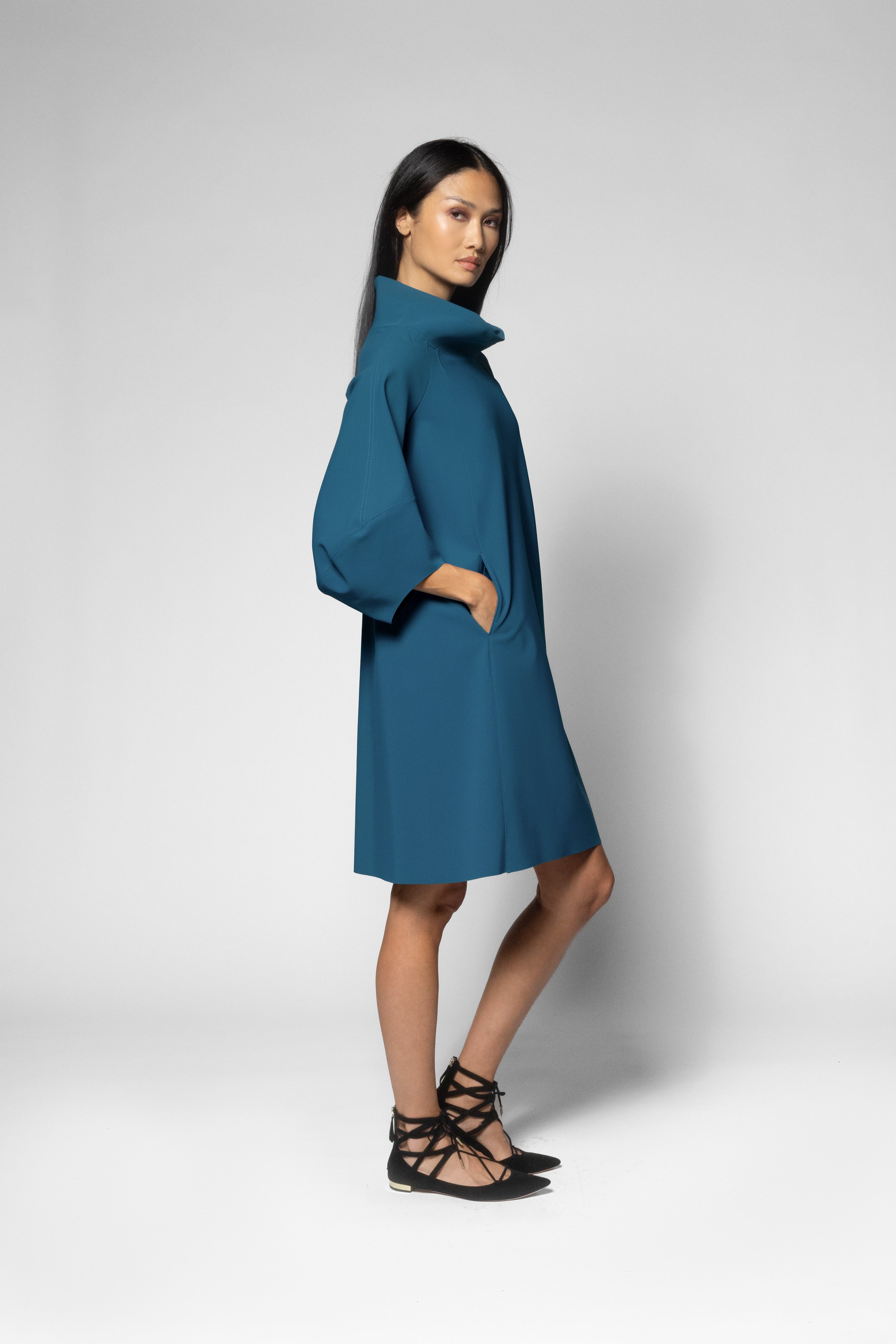 A woman modeling a teal blue high-neck, wide-sleeved shift dress with side pockets, paired with black lace-up strappy sandals, against a light gray studio background.