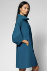 A woman modeling a teal blue turtleneck dress with wide draped sleeves and side pockets, photographed against a neutral light gray studio background.