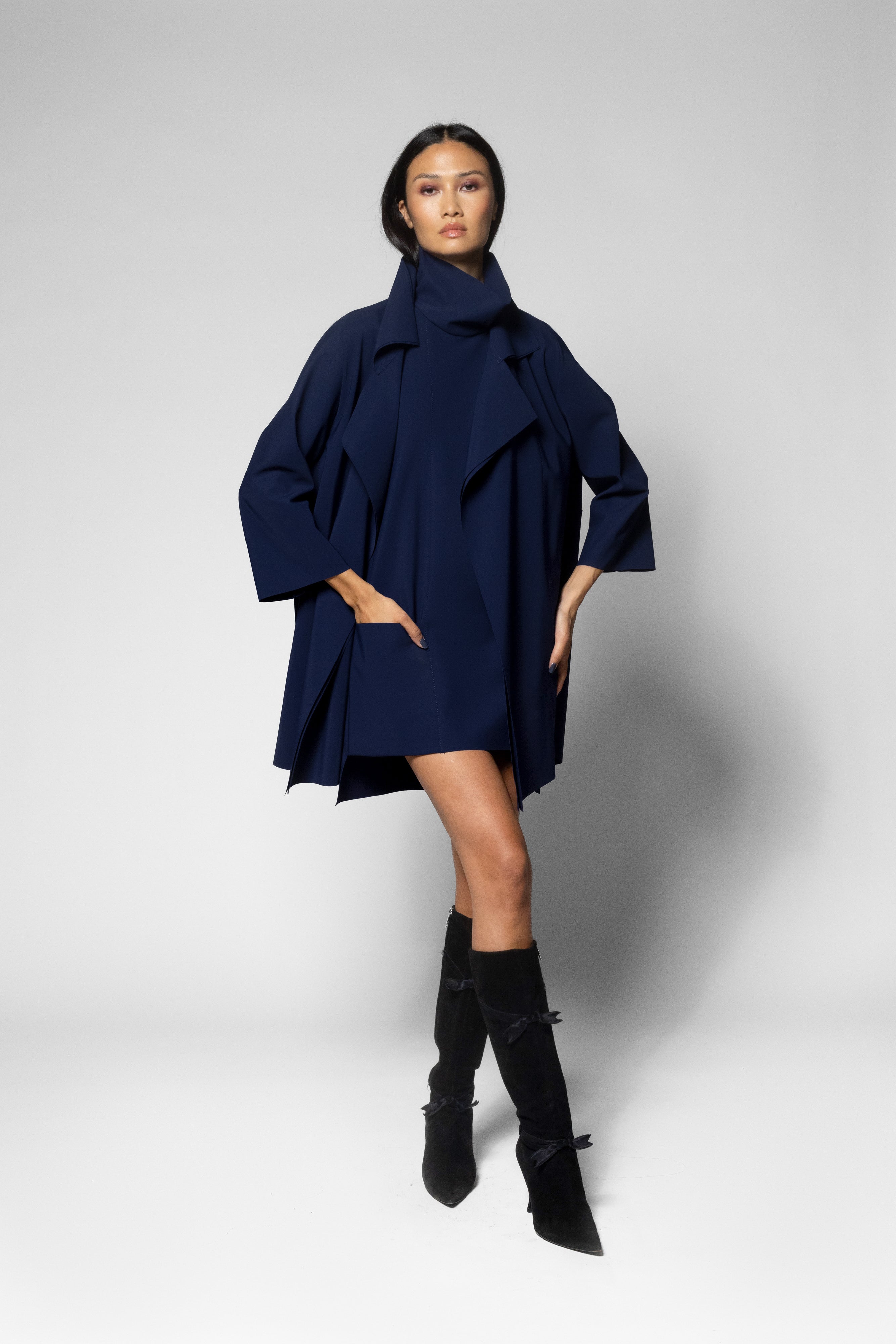 A model wearing a dark navy blue oversized coat with wide sleeves and a tunic with high collar, paired with black knee-high boots, against a light gray studio background.