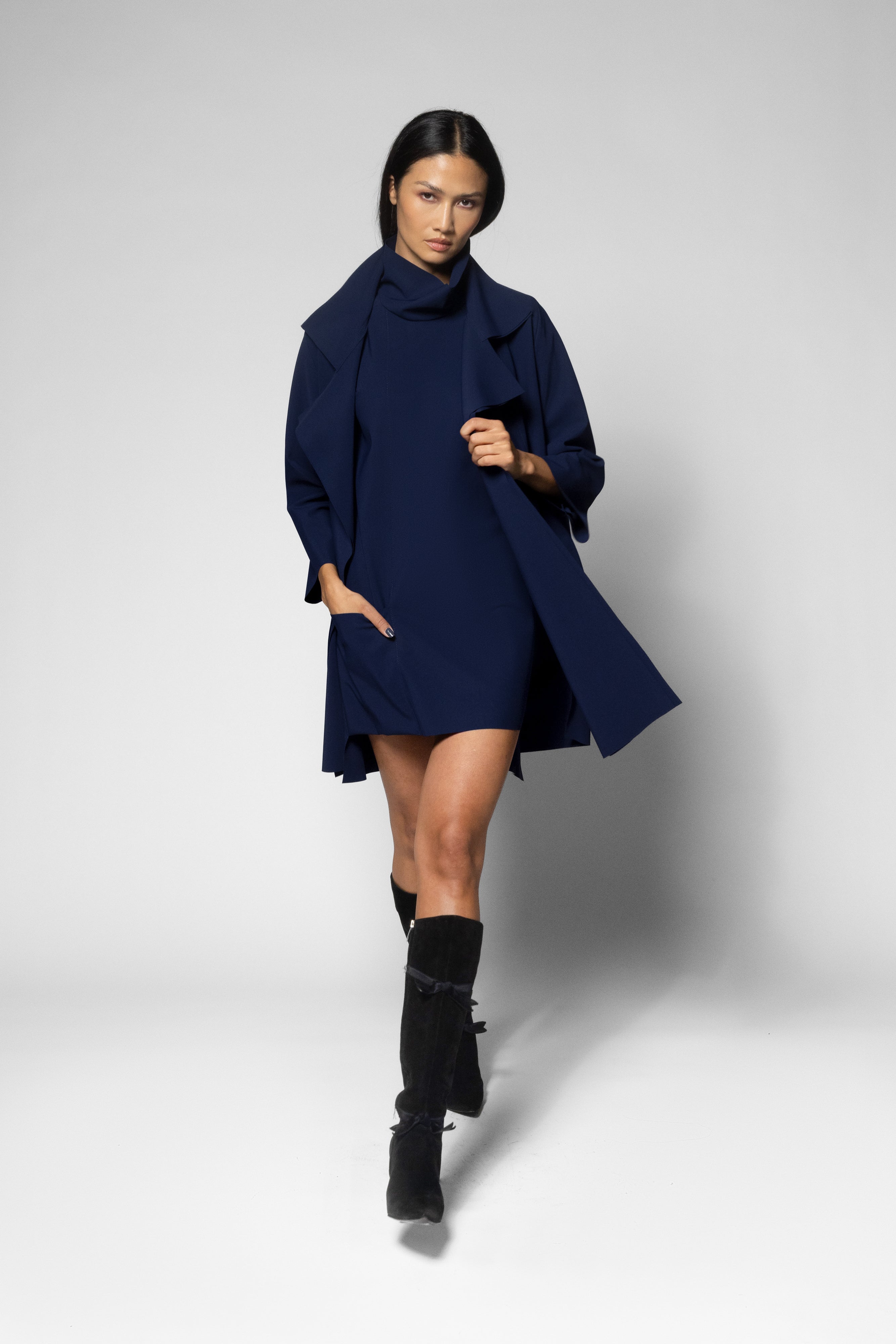 A model walks forward wearing a navy blue coat over a matching navy turtleneck dress, paired with black knee-high boots, against a light gray studio background.