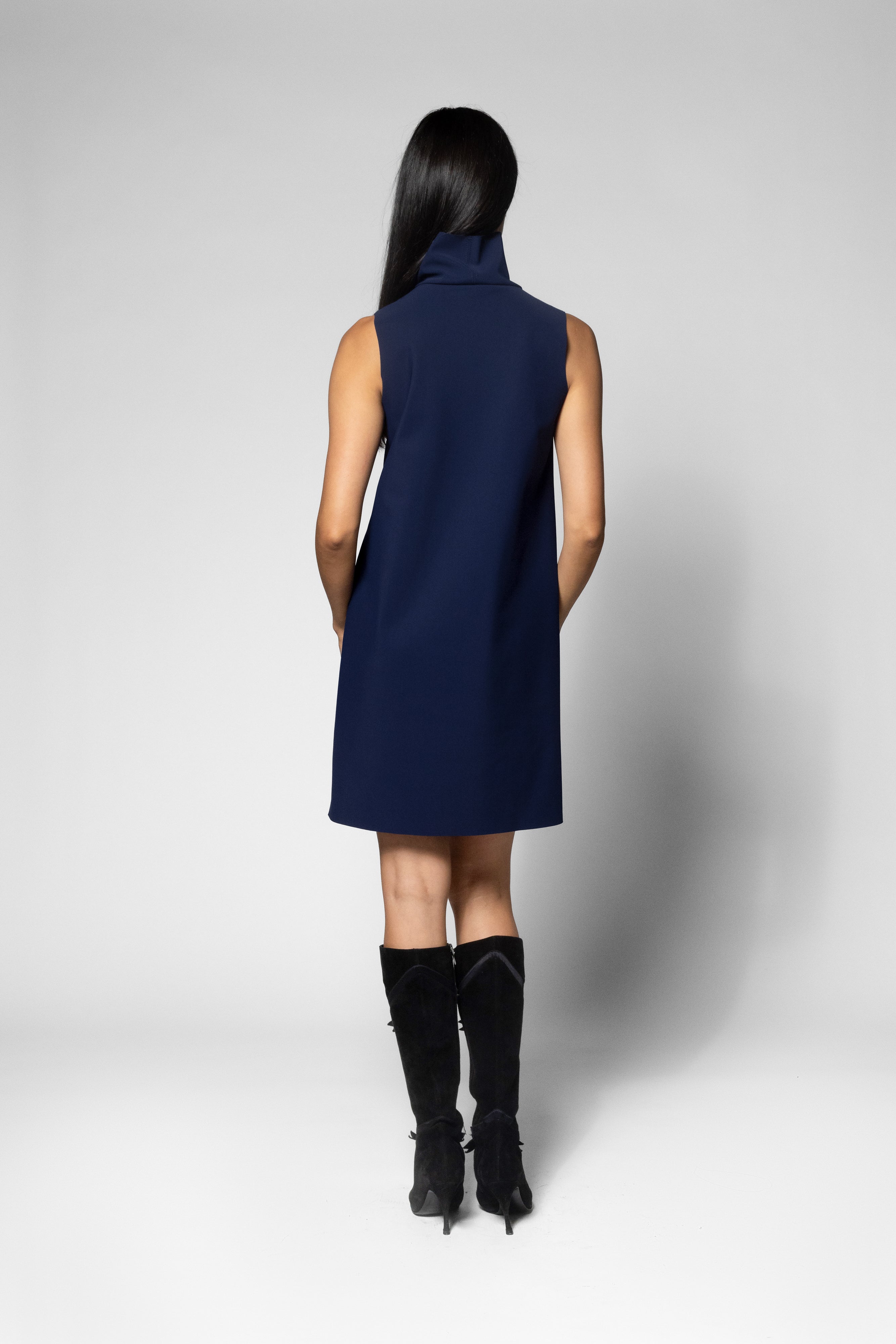 A woman shown from the back wearing a sleeveless navy blue high-neck dress and black knee-high boots, standing against a light gray studio background.