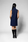 A woman shown from the back wearing a sleeveless navy blue high-neck dress and black knee-high boots, standing against a light gray studio background.