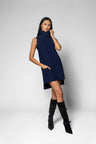 A woman modeling a sleeveless navy blue high-neck mini dress with side pockets, paired with black knee-high boots, against a light gray studio background.