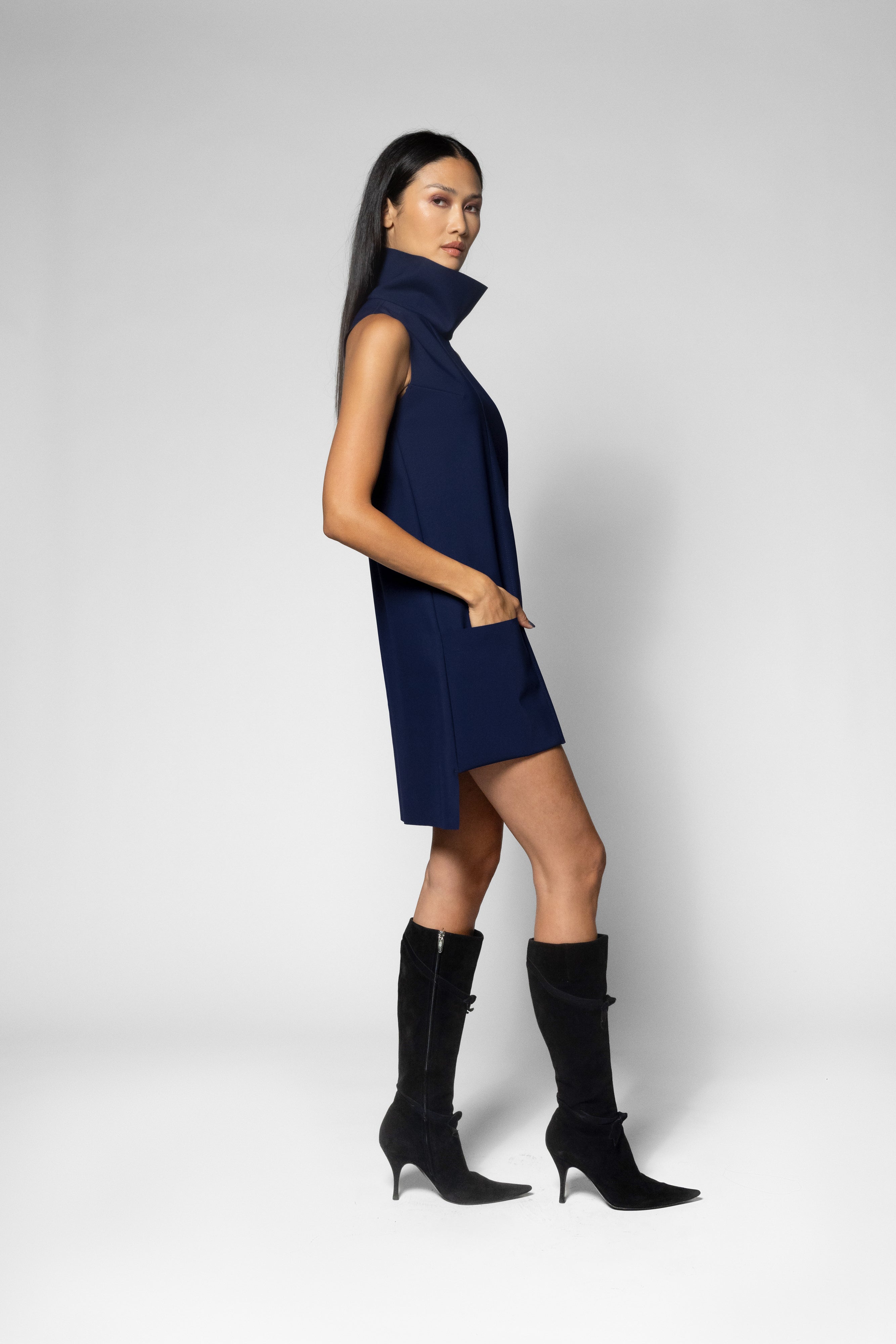 A woman modeling a sleeveless navy blue high-neck mini dress with black knee-high heeled boots, standing against a light gray studio background.