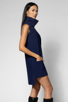 A woman modeling a sleeveless navy blue turtleneck mini dress with side pockets, paired with black boots, against a light gray studio background.