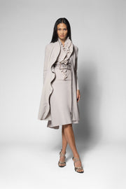 A model stands against a light gray studio background wearing a light beige ruffled wrap coat over a matching ruffled midi dress with a patterned belt, paired with beige strappy heels.