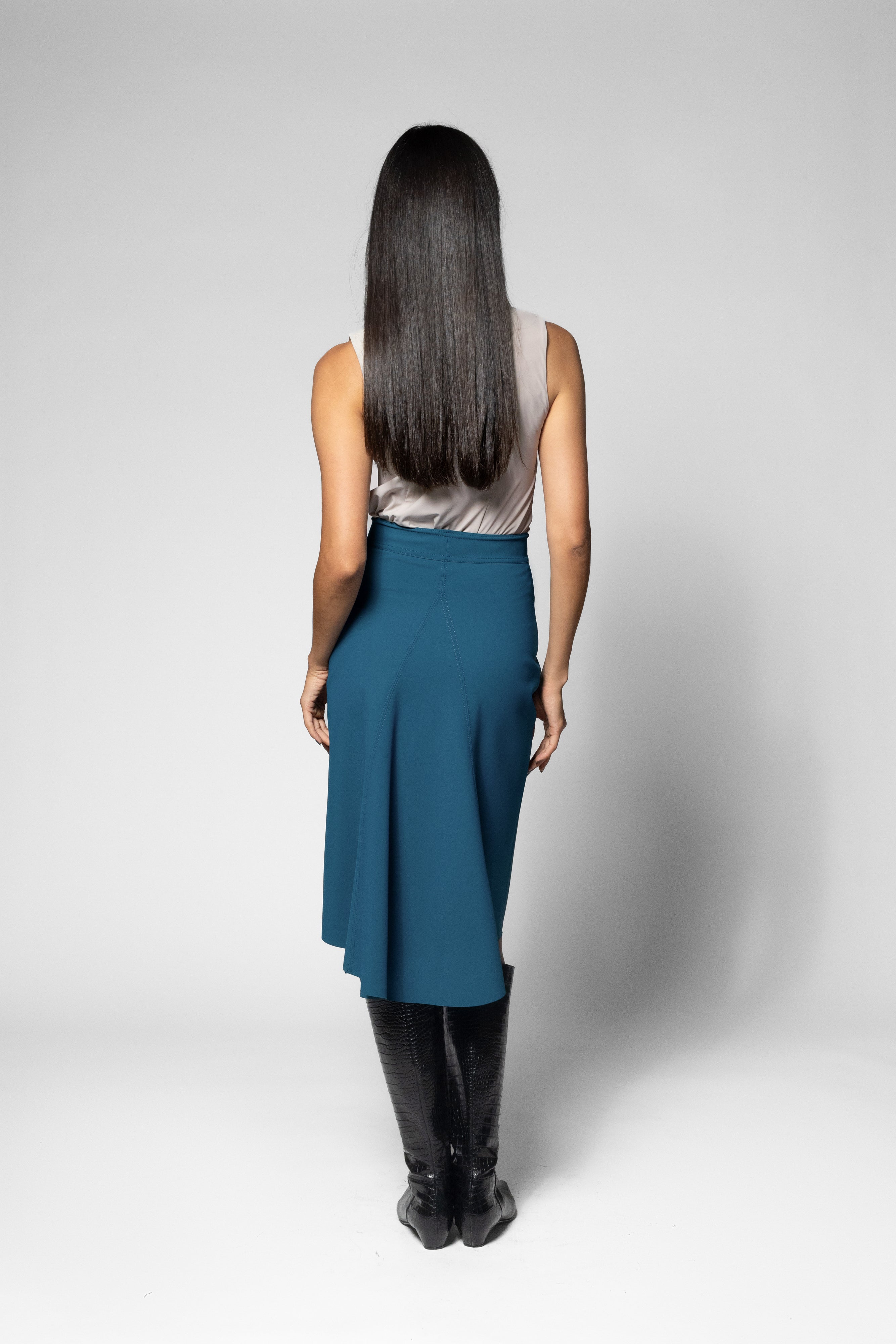 A woman viewed from behind wearing a sleeveless light gray top, a teal blue high-waisted skirt, and black knee-high boots, standing against a light gray studio background.