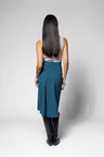 A woman viewed from behind wearing a sleeveless light gray top, a teal blue high-waisted skirt, and black knee-high boots, standing against a light gray studio background.
