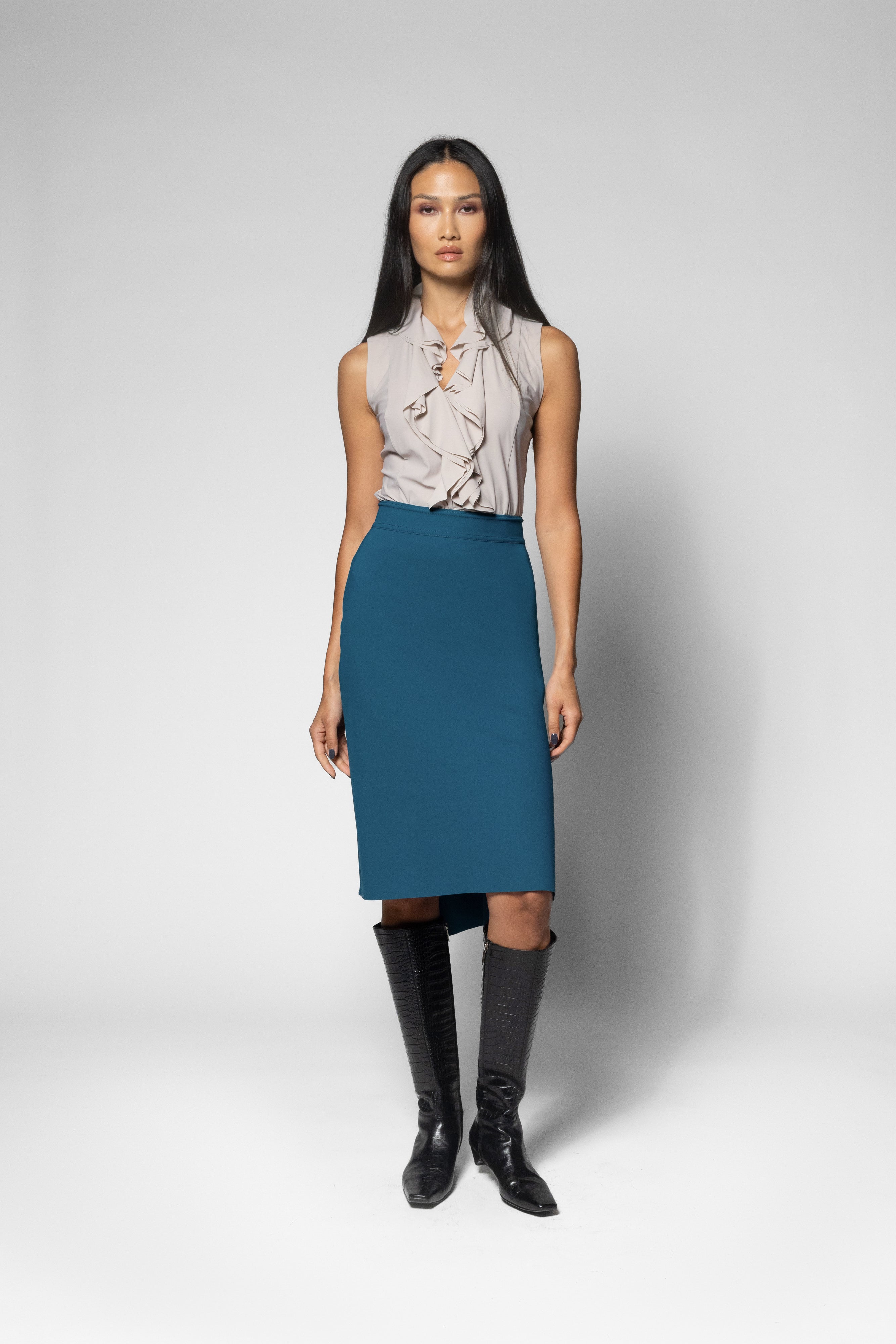 A model stands against a light gray studio background wearing a sleeveless light beige blouse with a ruffled tie-neck and a knee-length teal blue skirt, paired with black knee-high boots.