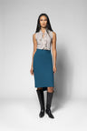 A model stands against a light gray studio background wearing a sleeveless light beige blouse with a ruffled tie-neck and a knee-length teal blue skirt, paired with black knee-high boots.