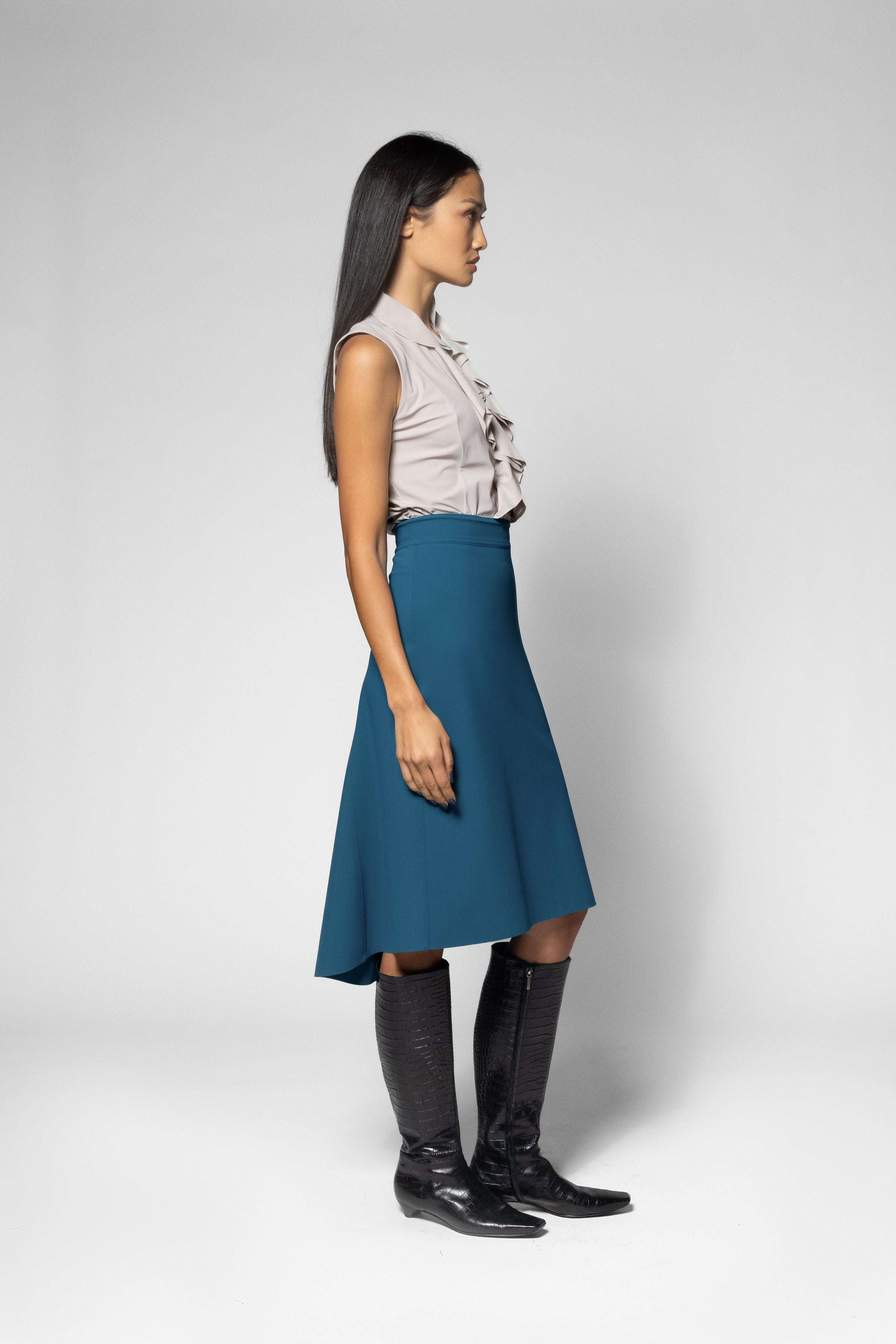 A woman stands in profile wearing a sleeveless light beige ruffled blouse, a teal A-line skirt, and black knee-high boots against a light gray studio background.