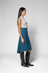A woman stands in profile wearing a sleeveless light beige ruffled blouse, a teal A-line skirt, and black knee-high boots against a light gray studio background.