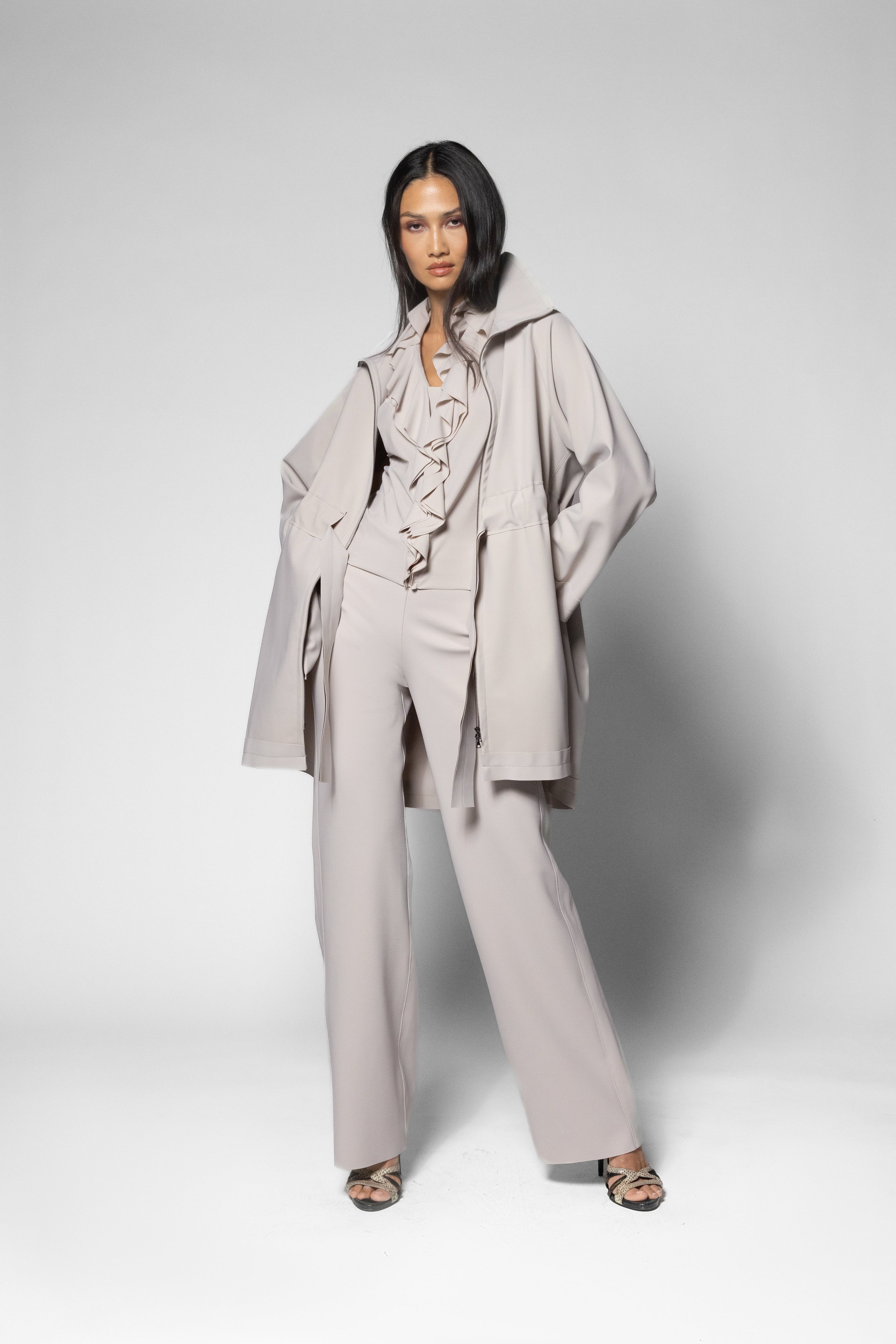 A model stands against a light gray studio background wearing a coordinated light taupe outfit consisting of a wide coat, pleated ruffled top, matching vest, and wide-leg trousers, paired with dark heeled sandals.