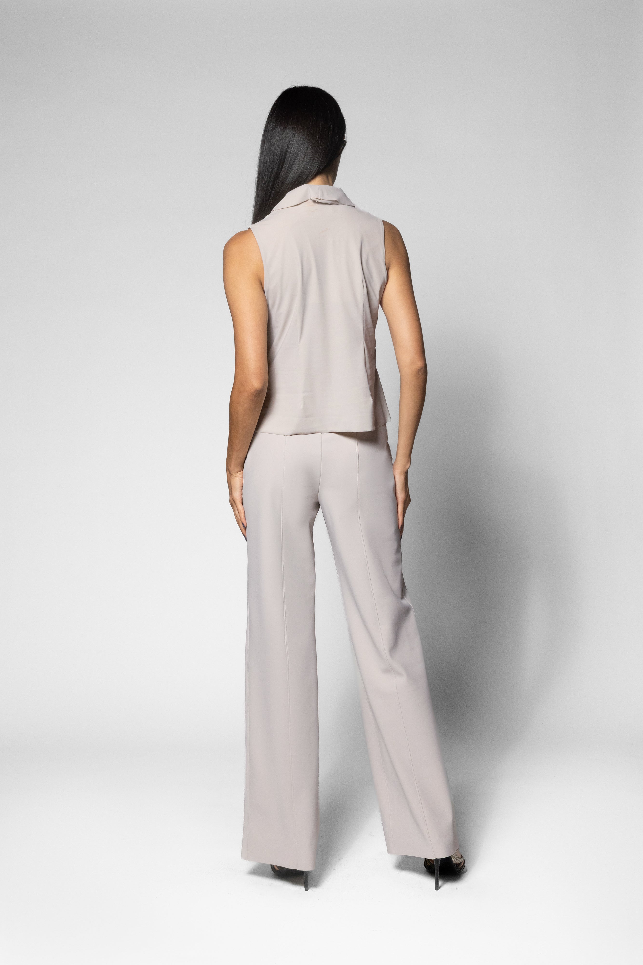 A woman is shown from the back wearing a light beige sleeveless collared top and matching wide-leg trousers, photographed against a neutral light gray studio background.