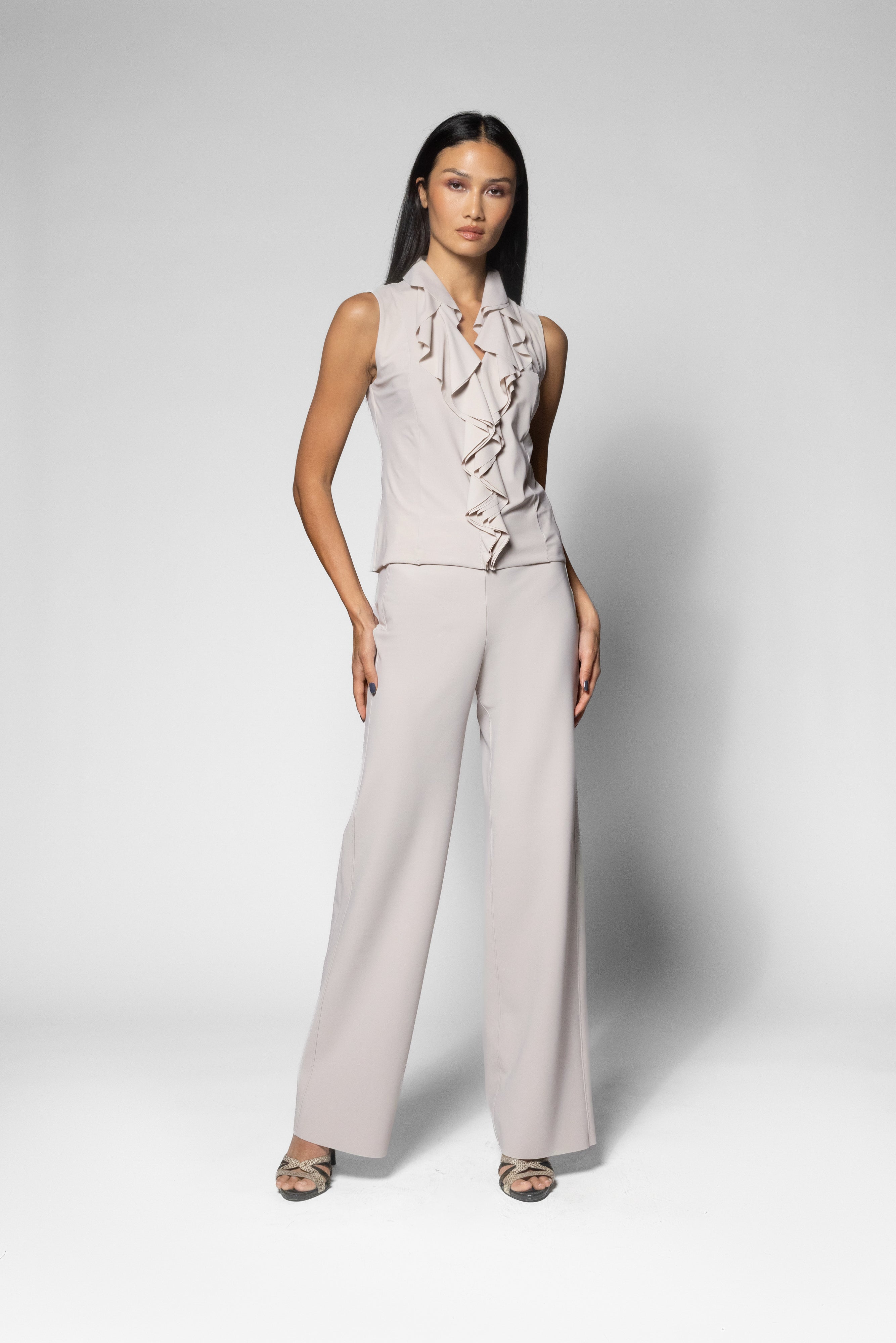 A woman modeling a sleeveless ruffled top and matching wide-leg trousers in a light blush pink or pale taupe color, standing against a light gray studio background.