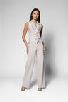 A woman modeling a sleeveless ruffled top and matching wide-leg trousers in a light blush pink or pale taupe color, standing against a light gray studio background.