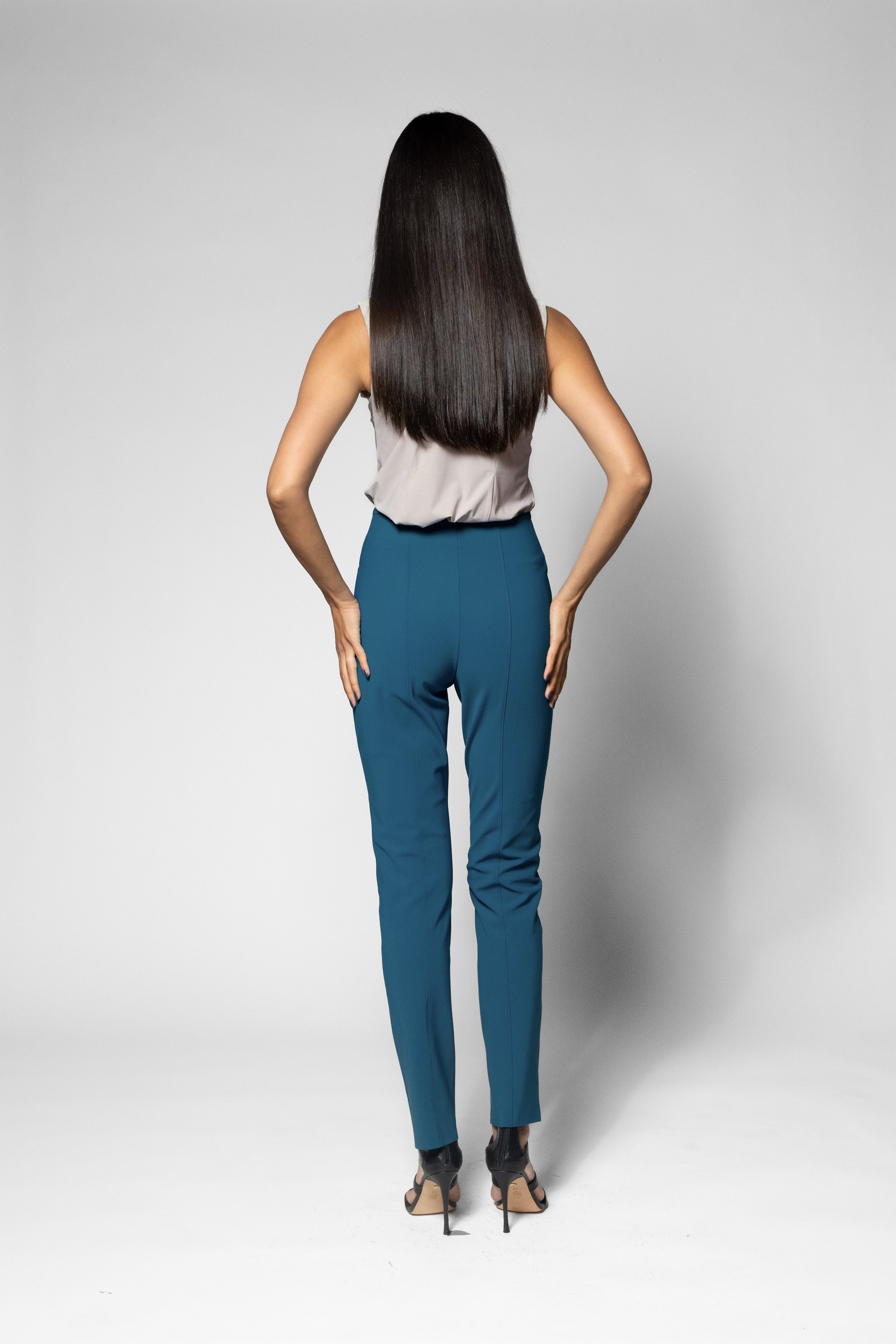 A woman with long dark hair is shown from behind wearing slim-fit teal pants, a light sleeveless top, and black high heels against a light gray studio background.