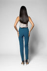 A woman with long dark hair is shown from behind wearing slim-fit teal pants, a light sleeveless top, and black high heels against a light gray studio background.