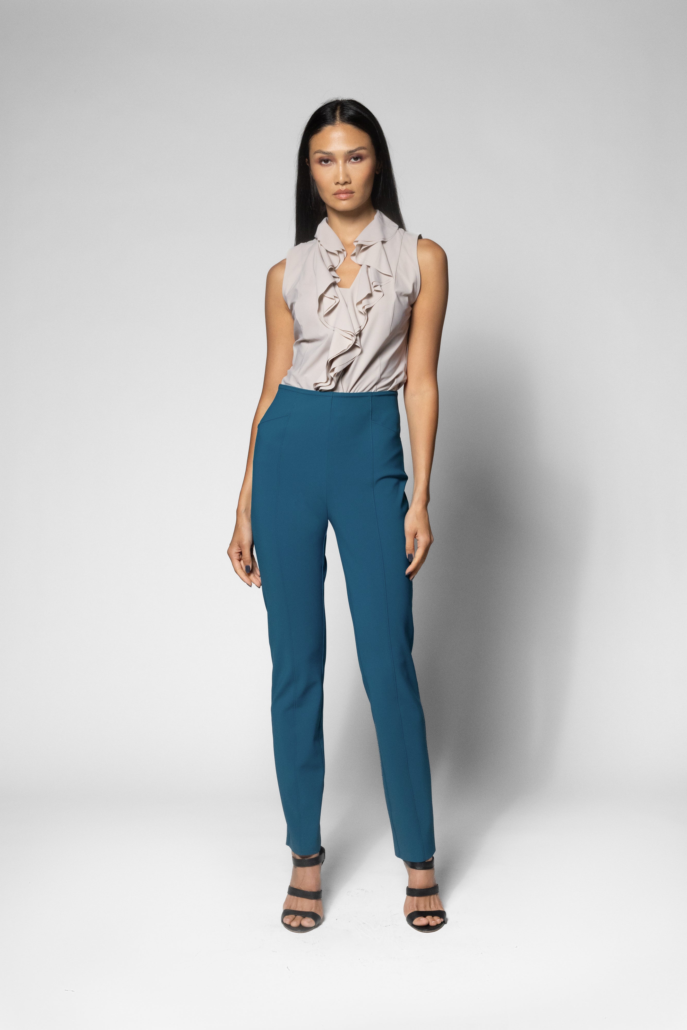 A woman modeling a sleeveless light beige ruffled blouse and high-waisted teal blue trousers, paired with black strappy heels, against a light gray studio background.