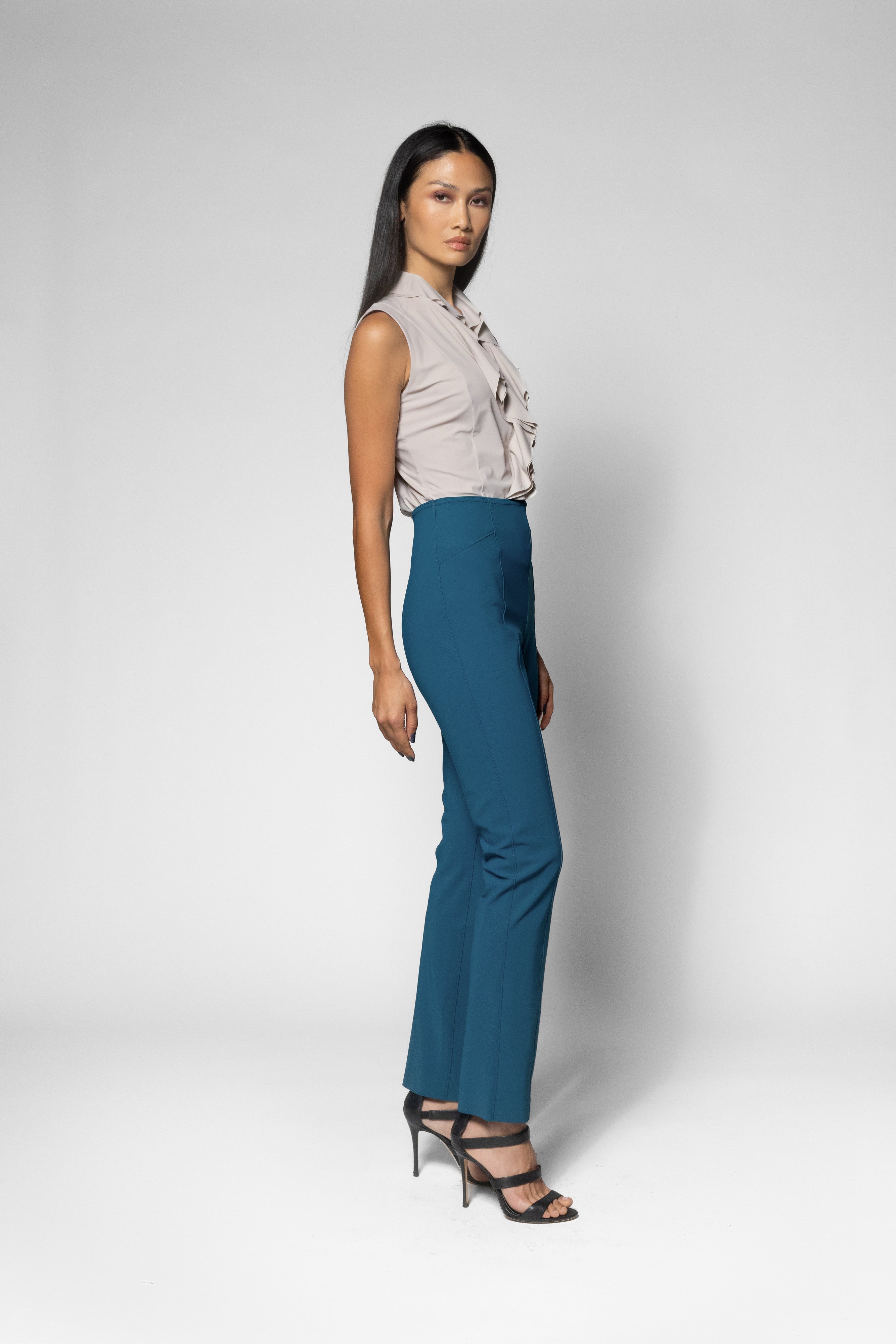 A woman modeling a sleeveless light beige top with ruffles and high-waisted teal blue wide-leg trousers, paired with black strappy heels, against a light gray studio background.