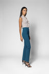 A woman modeling a sleeveless light beige top with ruffles and high-waisted teal blue wide-leg trousers, paired with black strappy heels, against a light gray studio background.