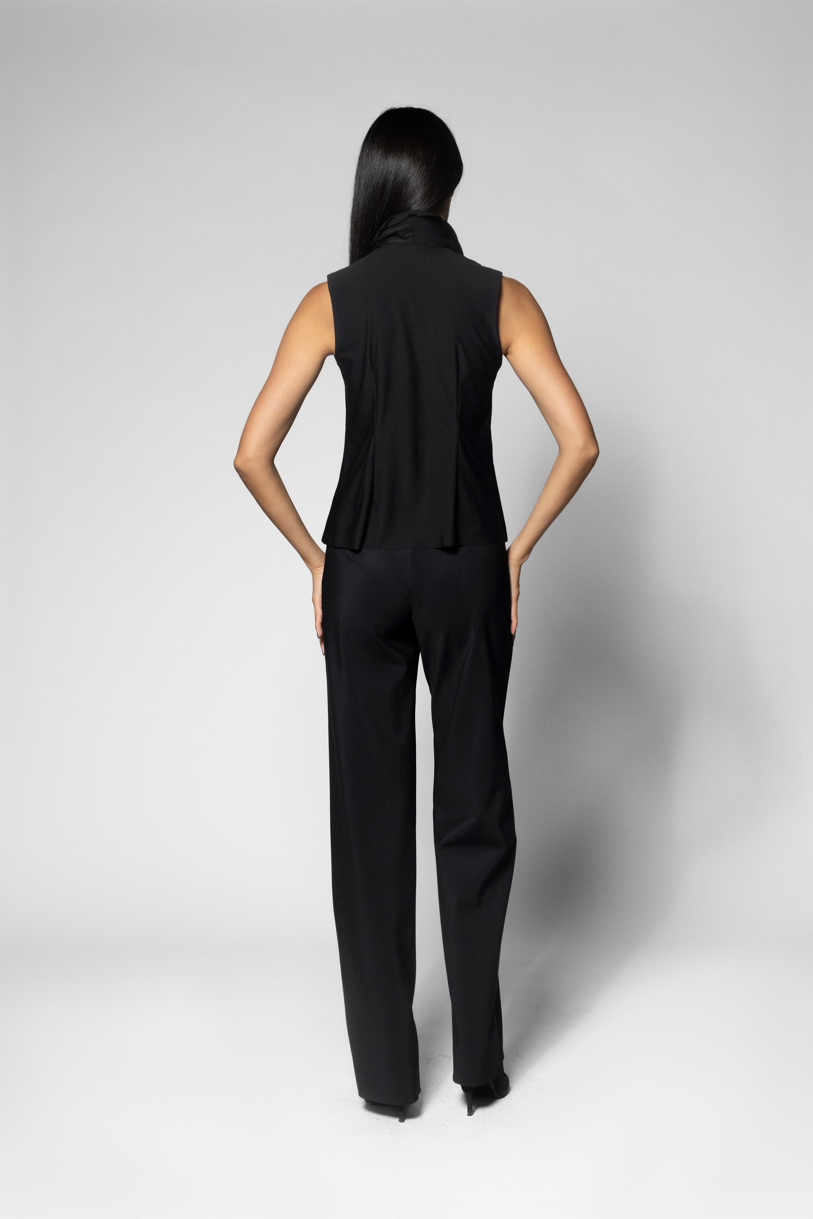 A woman shown from the back wearing a black sleeveless high-neck top and matching black wide-leg trousers against a light gray studio background.