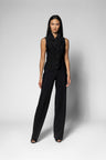 A model stands against a light gray studio background wearing a black sleeveless top with a ruffled collar and matching black wide-leg pants.