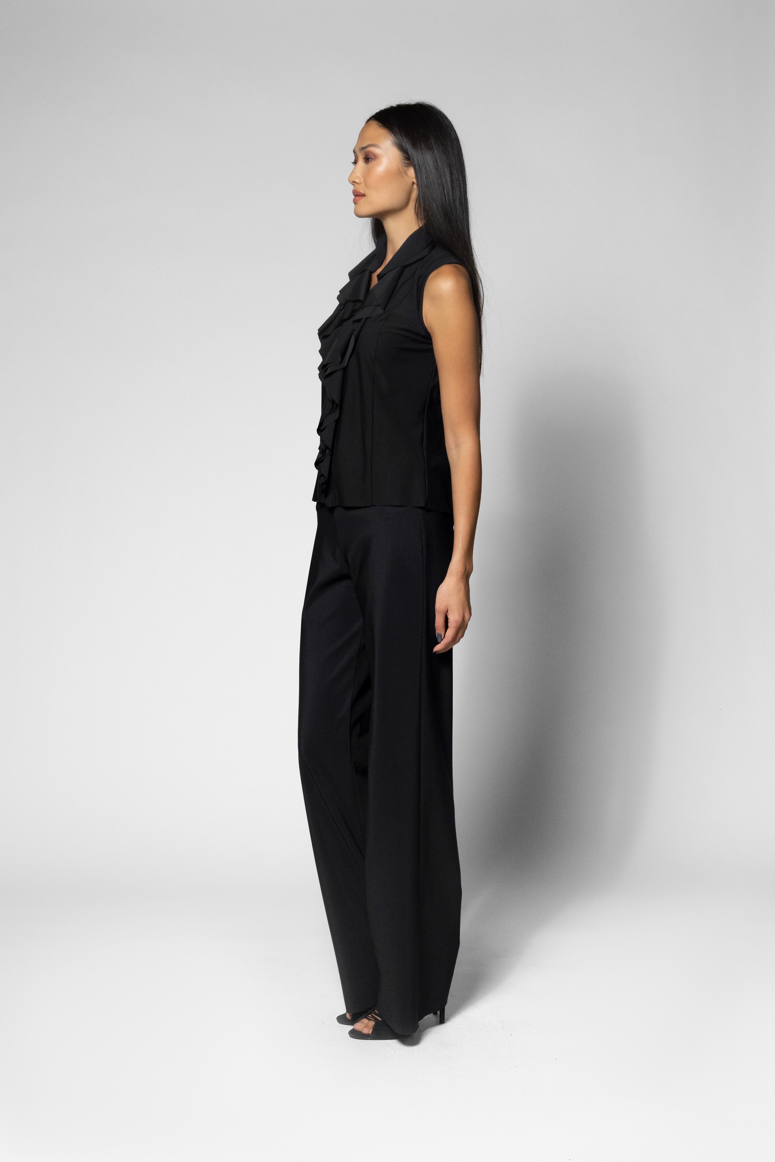 A woman stands in profile wearing a black sleeveless top with ruffled detailing and matching black wide-leg pants against a light gray studio background.