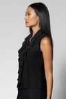 A woman stands in profile wearing a black sleeveless top with ruffled detailing and matching black wide-leg pants against a light gray studio background.