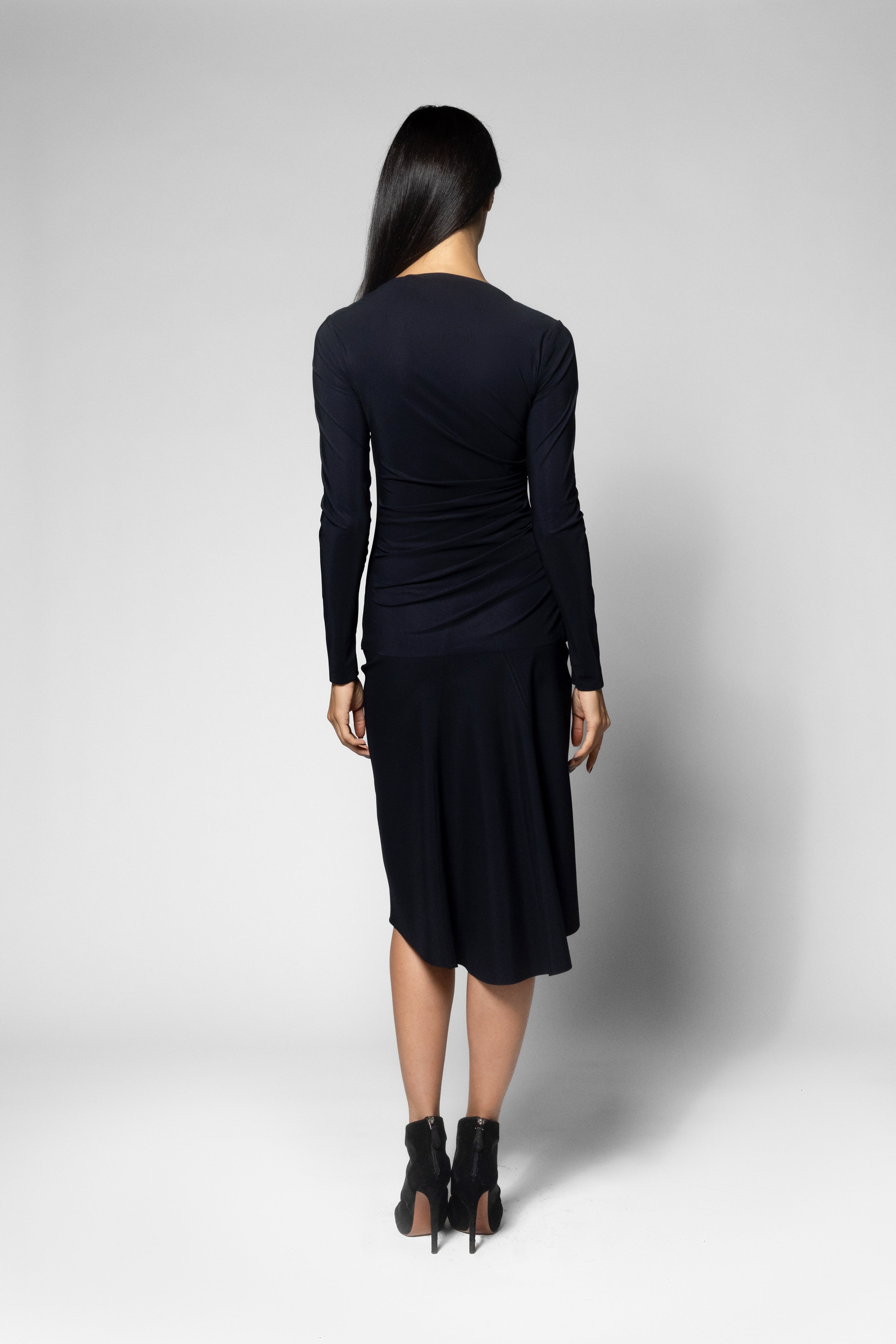 A woman viewed from the back wearing a dark navy long-sleeve top with gathered draping at the waist and black pointed-toe heels, photographed against a light gray studio background.