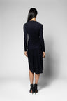 A woman viewed from the back wearing a dark navy long-sleeve top with gathered draping at the waist and black pointed-toe heels, photographed against a light gray studio background.